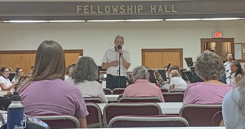 Pekin Park Concert Band Director Steve Skinner welcomes the assembled crowd as they settle in for an evening of music inside the Fellowship Hall of the United Methodist Church, a last-minute weather-safe haven from the summer heat.